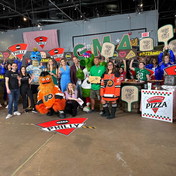 A group of people and mascots pose with "GMA Philly" and "Pizza Jawn" signs.