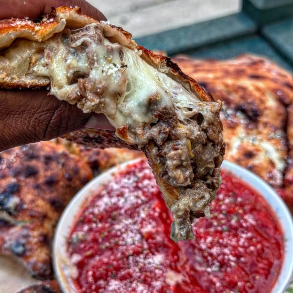 A hand holds a cheesy, meaty calzone slice over a bowl of red marinara sauce.