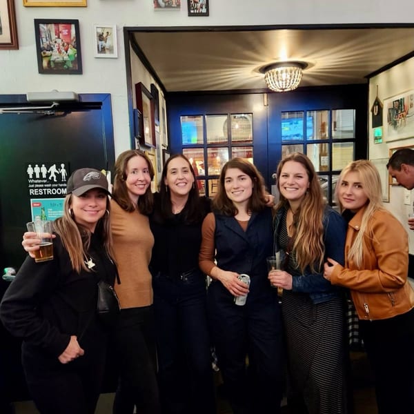 Six women smiling and holding drinks inside Bar Jawn, with a restroom sign visible.