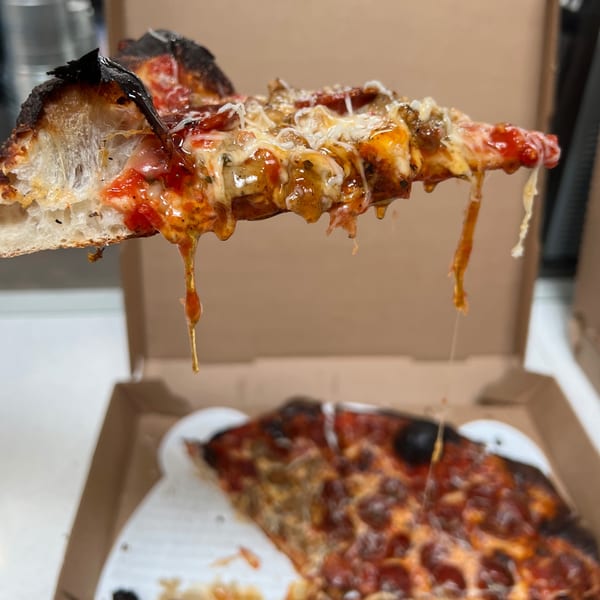 A slice of Bar Jawn pizza with melted cheese and toppings dripping, held above a pizza box.