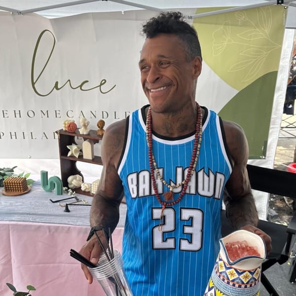 A smiling man in a blue Bar Jawn jersey holds a pitcher of ice and a cup of straws.