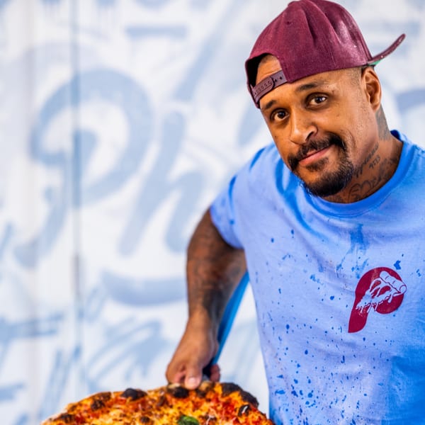 A man in a blue shirt and maroon cap holds a Bar Jawn pizza, smiling at the camera.
