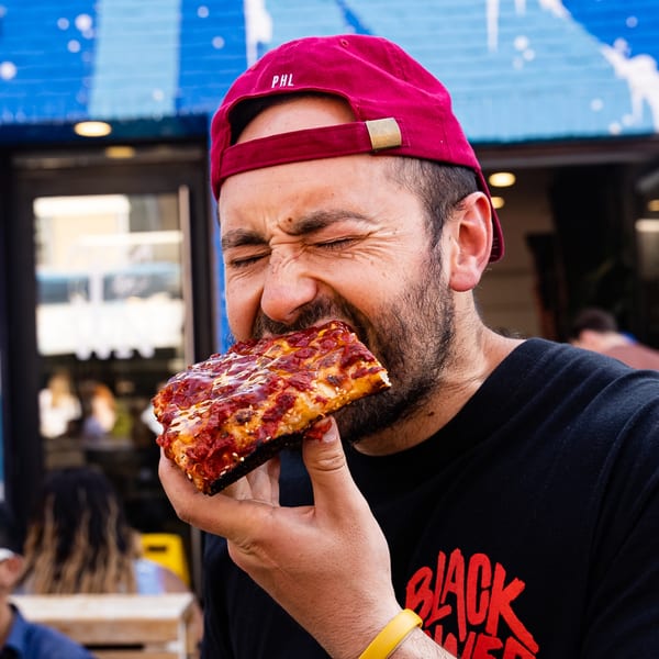 Man in a red cap intensely bites into a square slice of Bar Jawn pizza outdoors.