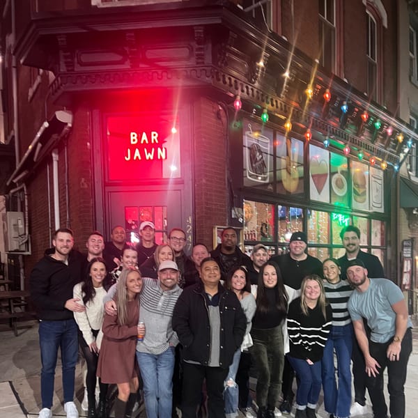 A large group of smiling people gathered outside Bar Jawn at night, illuminated by a red neon sign.