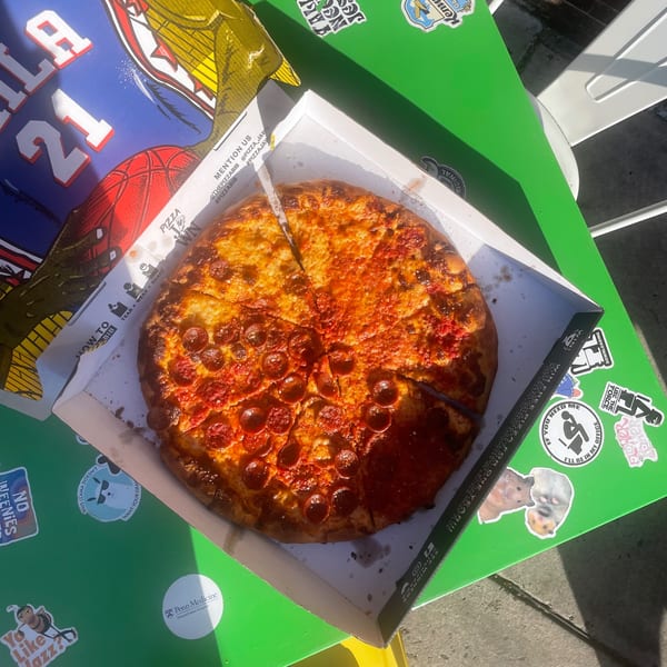 A pepperoni pizza from Bar Jawn in a box on a green table with stickers, next to a basketball jersey.