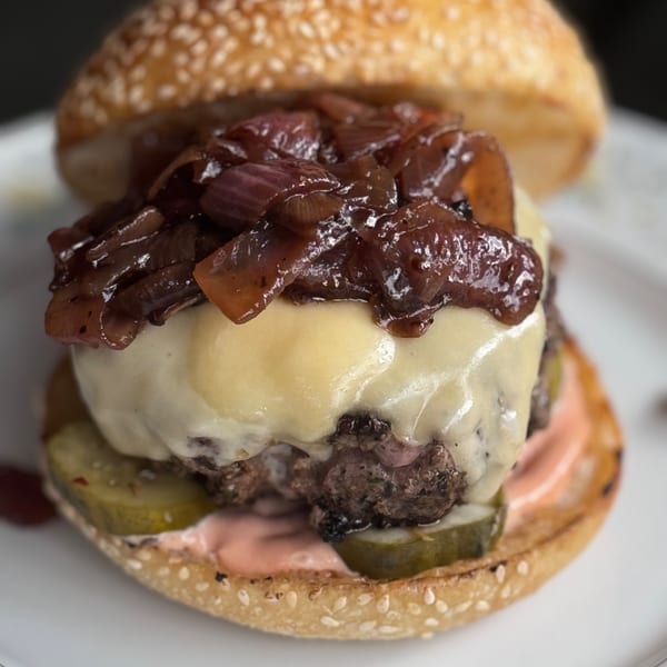 Close-up of a Bar Jawn burger with melted cheese, caramelized onions, pickles, and sauce on a sesame bun.