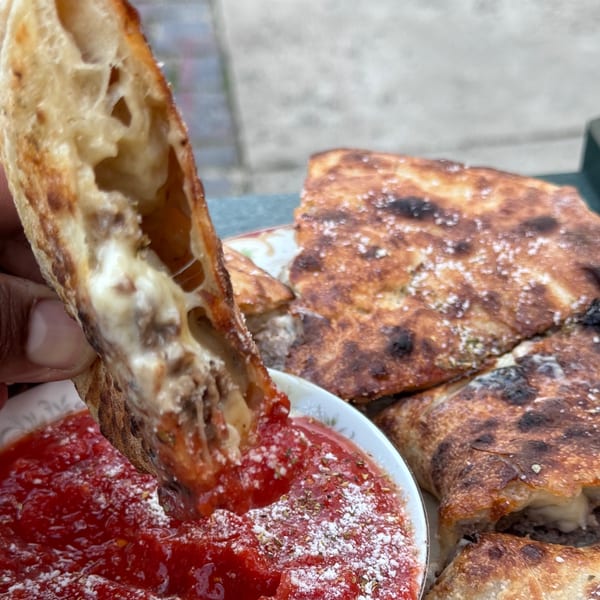 A hand dips a cheesy, crusty slice of Bar Jawn pizza into a bowl of red sauce.