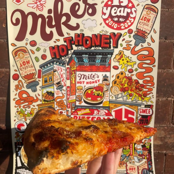 A hand holds a slice of Bar Jawn's pizza in front of a colorful Mike's Hot Honey poster.